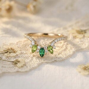 Könnte beinhalten: Ein goldfarbener Ring mit einem gebogenen Band, besetzt mit kleinen Diamanten. Der Ring hat einen zentralen smaragdgrünen Edelstein, flankiert von zwei birnenförmigen grünen Edelsteinen und kleineren Diamanten, die ein blattartiges Design bilden. Der Ring liegt auf einer weißen Spitzentischdecke.