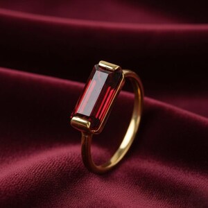 Könnte beinhalten: Ein goldener Ring mit einem rechteckigen, tiefroten Edelstein. Der Edelstein ist in einem goldenen Band mit schlichtem Design gefasst. Der Ring wird vor einem bordeauxfarbenen Hintergrund gezeigt.