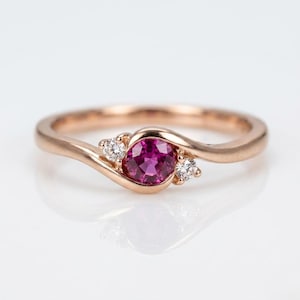 Könnte beinhalten: Roségoldener Ring mit einem leuchtenden magentafarbenen Edelstein in der Mitte, flankiert von zwei kleineren, funkelnden weißen Steinen. Das Band hat ein fließendes, elegantes Design. Der Ring ist vor einem hellen, reflektierenden Hintergrund platziert.