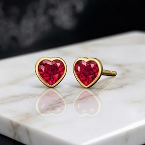 Könnte beinhalten: Ein Paar herzförmige Ohrstecker. Jeder Ohrring hat einen leuchtend roten Edelstein, der in einer goldfarbenen Lünette gefasst ist. Die Ohrringe werden auf einer weißen Marmoroberfläche präsentiert, die ihre Form widerspiegelt. Romantischer Schmuck.