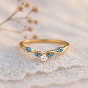 Könnte beinhalten: Ein goldfarbener Ring mit geschwungenem Design. Der Ring hat kleine, runde, türkisfarbene Steine und einen einzelnen, klaren, runden Stein in der Mitte. Der Ring ist vor einem weichen, weißen Hintergrund platziert.