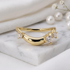 Può includere: Un anello d'oro curvo con tre pietre preziose trasparenti. L'anello ha una finitura liscia e lucida ed è posto su una superficie di marmo bianco. Le pietre preziose sono rotonde e scintillanti, aggiungendo un tocco di eleganza.