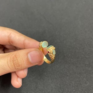 May include: A gold-colored ring with an open design, featuring two oval, light green gemstones and a small red gemstone. The ring has an ornate, textured band and is held against a dark background.