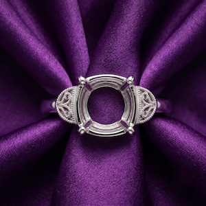 Könnte beinhalten: Eine Ringfassung aus silberfarbenem Metall mit einer zentralen, kreisförmigen Krappenfassung für einen Edelstein. Die Ringschultern weisen filigrane Ausschnitte und ein perlenartiges Milgrain-Detail entlang der Metallkante auf.