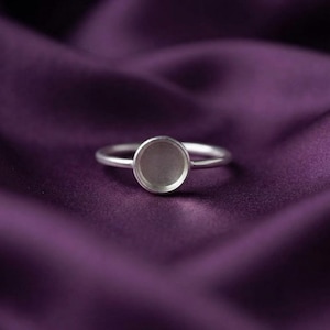 Könnte beinhalten: Ein silberfarbener Ring mit einer schlichten, kreisförmigen Fassung. Der Ring hat eine glatte, dünne Ringschiene und eine flache, leere kreisförmige Vertiefung in der Mitte, die für einen Stein oder Cabochon vorgesehen ist. Das Metall ist matt gebürstet.