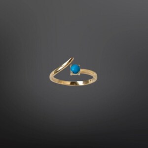 Könnte beinhalten: Ein goldfarbener Ring mit einem leuchtend türkisfarbenen Stein. Der Ring hat ein einzigartiges offenes Design mit einem gebogenen Band, das sich um den Finger legt, und einem kleinen, quadratisch gefassten Türkis-Edelstein.