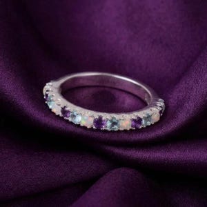 Könnte beinhalten: Ein silberfarbener Ring mit einer Reihe abwechselnd gesetzter, runder Edelsteine. Das Design umfasst irisierende weiße Opale, tiefviolette Amethyste und hellblaue, aquamarinfarbene Steine, die in einer durchgehenden Fassung im Metallring sitzen.