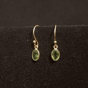 Könnte beinhalten: Ein Paar goldfarbene Ohrhänger. Jeder Ohrring hat einen kleinen, oval geformten, hellgrünen Edelstein, der an einem gebogenen Haken hängt. Die Ohrringe werden vor einem dunklen Hintergrund präsentiert.
