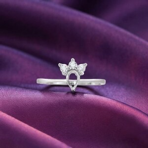 Könnte beinhalten: Ein silberfarbener Ring mit einer kronenförmigen Fassung, die drei kleine, klare, rund geschliffene Steine in horizontaler Anordnung enthält. Die Mitte des Kronendesigns weist eine tropfenförmige Öffnung auf und die Ringschiene ist schmal.