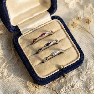 Könnte beinhalten: Drei silberne Ringe mit Edelsteinakzenten werden in einer blauen Samt-Schmuckschatulle präsentiert. Jeder Ring hat ein einzigartiges Design mit roten, blauen und hellblauen Edelsteinen. Die Schatulle ist geöffnet und zeigt die Ringe.