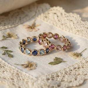Könnte beinhalten: Zwei goldene Ringe mit farbigen Edelsteinen. Ein Ring hat blaue und hellblaue Steine, der andere rosa Steine. Die Ringe liegen auf einem weißen, mit Spitze besetzten Tuch, mit getrockneten Blumen verstreut.