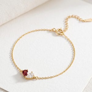 Könnte beinhalten: Ein zartes, goldfarbenes Armband mit zwei tropfenförmigen Edelsteinen: einem roten und einem klaren. Das Armband hat eine feine Kette und einen verstellbaren Verschluss. Die Edelsteine sind eng beieinander gesetzt, was ein subtiles, elegantes Design ergibt.