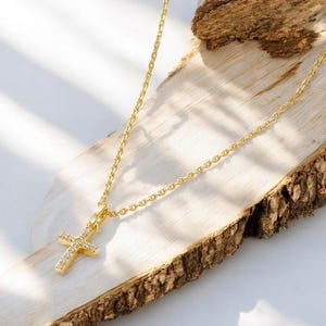Può includere: Una collana dorata con un pendente a forma di croce ornato di piccole pietre preziose trasparenti. La delicata catena poggia su una superficie di legno su uno sfondo chiaro. Il pendente a croce misura circa 2 cm di lunghezza. Un gioiello.