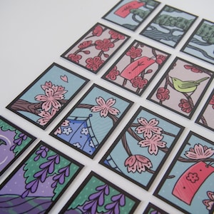 Pode incluir: Um conjunto de cartões retangulares com ilustrações de inspiração japonesa. Cada cartão apresenta uma borda preta e um design único, incluindo flores de cerejeira, pássaros e paisagens em cores pastel. Os cartões estão dispostos em uma superfície branca.