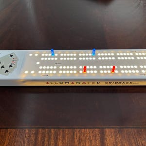 May include: Illuminated cribbage board with blue and red pegs. The board has a white surface with rows of illuminated holes. The text "ILLUMINATED CRIBBAGE.com" is printed on the board. A deck of cards is shown on the left side.