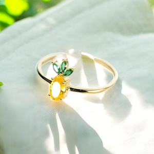 Könnte beinhalten: Ein goldfarbener Ring mit Ananas-Design. Die Ananas hat eine gelbe, ovale Mitte und grüne Blätter. Der Ring ist hinten offen und liegt auf einem weißen Tuch. Das Wort "POMELA" und der Satz "subtle. refined. unique." befinden sich oben links.