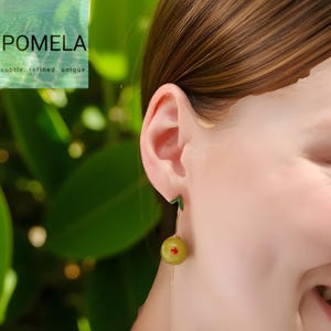 Op de afbeelding: Olijfvormige oorbellen met een rode accent, hangend aan een goudkleurige draad. De oorbellen hebben een groen blad detail aan de bovenkant. De afbeelding bevat de tekst "POMELA" en "subtle. refined. unique."