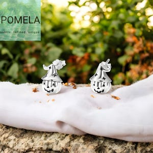 Puede incluir: Dos pendientes plateados con temática de Halloween. Cada pendiente presenta un diseño de calabaza con un sombrero de bruja. Las calabazas tienen caras talladas. Los pendientes están sobre una tela blanca con un fondo verde borroso. La marca "POMELA" es visible.