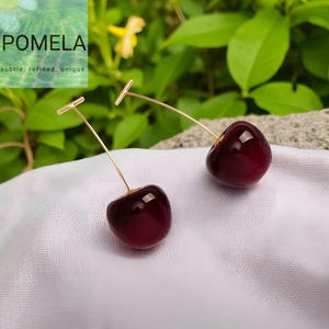May include: A pair of cherry-shaped earrings. Each earring has a deep red, glossy cherry suspended from a delicate gold-coloured wire. The earrings rest on white fabric, with a blurred green background.