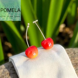 May include: A pair of cherry earrings with red and yellow gradient fruit-shaped pendants. The earrings have long, thin, silver-colored stems. The earrings are displayed on a white textured surface, with green foliage in the background.