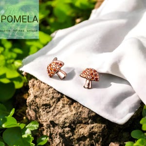 以下が含まれることがあります： ローズゴールド仕上げとオレンジ色の宝石のキャップが付いた、キノコ型のカフスボタン2組。カフスボタンは白い布の上に展示されています。「POMELA」のブランド名と「subtle. refined. unique.」というキャッチコピー。