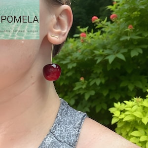 May include: A pair of gold-toned earrings featuring a cherry design. The earrings have a long, slender gold wire that suspends a large, glossy, dark red cherry. The earrings are shown against a background of green foliage.