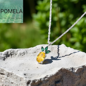 Op de afbeelding: Een zilveren ketting met een ananas hanger. De hanger heeft een gele ovale edelsteen en groene bladeren. De ketting wordt getoond op een getextureerd, lichtgrijs oppervlak met een groene achtergrond.