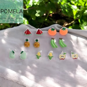 May include: A selection of fruit-shaped earrings, featuring watermelon slices, oranges, pineapples, bananas, pears, sweetcorn, and apples. The earrings are brightly coloured and detailed, with silver posts. The brand name "POMELA" is visible in the upper left corner.