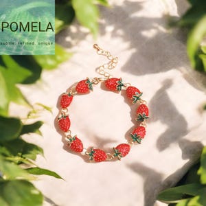 May include: A gold-toned bracelet featuring red strawberry charms with green leaves. The bracelet is adjustable and has the word "POMELA" and the text "subtle. refined. unique." in the upper left corner.