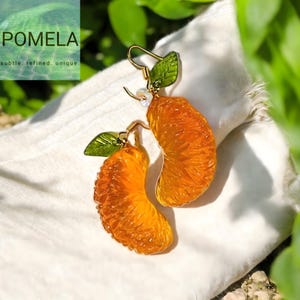 Puede incluir: Pendientes con forma de gajos de naranja, con ganchos dorados y detalles de hojas verdes. Los pendientes se muestran sobre un fondo blanco. El texto "POMELA" y las palabras "subtle. refined. unique." son visibles.