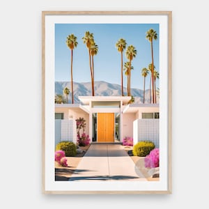 Op de afbeelding: Een wit modern huis uit het midden van de eeuw met een feloranje deur, omringd door palmbomen en roze bloemen. Het huis heeft een witte bakstenen buitenkant en een plat dak. De deur bevindt zich in het midden aan de voorkant van het huis en is omgeven door een wit frame. De palmbomen zijn hoog en slank, en de bloemen bloeien. Het huis ligt op de achtergrond van bergen.