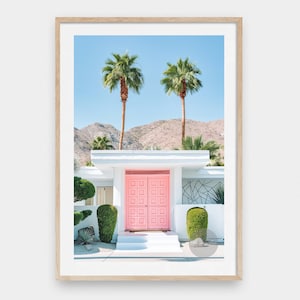 Op de afbeelding: Een wit modern huis uit het midden van de eeuw met een felroze dubbele deur, omringd door palmbomen en een woestijnlandschap.