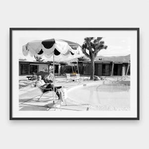 May include: Black and white framed print of a mid-century modern scene. A person sits in a chair under a large, scalloped umbrella near a pool. The image captures a retro aesthetic with a focus on leisure and architecture.