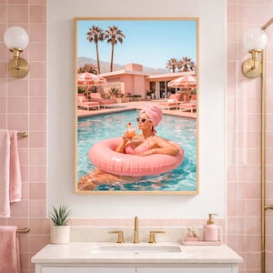 May include: A framed print depicts a woman in a pink swimsuit and towel, floating in a pool with a pink inflatable ring. The scene includes a pink resort, palm trees, and a clear blue sky, creating a retro, vacation theme.