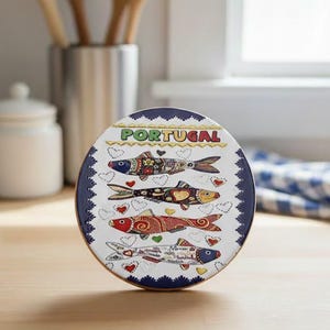 Può includere: Un piatto decorativo rotondo e bianco con un bordo blu con la scritta "PORTOGALLO" e quattro illustrazioni di pesci colorati. Ogni pesce ha motivi unici e accenti a forma di cuore. Il piatto è su una superficie di legno.