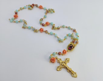 May include: A rosary with light blue, brown, and peach-colored beads, connected by gold-colored wire. Peach-colored rose-shaped beads are interspersed throughout. The rosary ends with a gold-colored crucifix and a small oval pendant with a religious image.