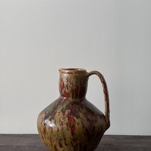 Op de afbeelding: Een keramische kan met een afgeronde basis en een handvat. De kan heeft een gespikkelde glazuur in tinten bruin, groen en rood. De kan staat op een donker houten oppervlak.