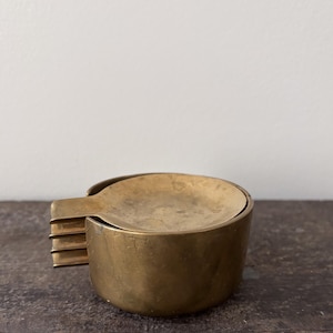 May include: A circular brass ashtray with a smooth, polished finish. The object features a wide, flat rim and a distinct side extension with three horizontal, tiered ridges that protrude from the main cylindrical body of the metal container.