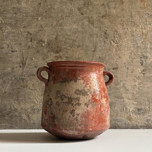 Op de afbeelding: Een terracotta pot met twee zijhandvatten, voorzien van een verweerde, rustieke afwerking met plekken van blootliggende klei en een vervaagde roodbruine glazuurlaag. De pot heeft een brede, ronde vorm en een uitlopende rand met textuur.