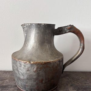 May include: A rustic metal pitcher with a hammered texture and a weathered, dark grey finish. The vessel features a wide, rounded base, a tapered neck, a small pouring spout, and a curved handle with visible copper-toned oxidation and wear.