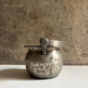 Op de afbeelding: Een kleine, ronde metalen pot met een doffe grijze afwerking en een getextureerd, verweerd oppervlak. Het heeft een enkele, dikke, scharnierende handgreep die aan de rand is bevestigd met een cirkelvormig, decoratief klinknageldetail.