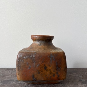 May include: A square-shaped ceramic vase with a short, rounded neck and a wide opening. The surface features a mottled, textured glaze in shades of burnt orange, deep brown, and charcoal gray, creating a weathered appearance across the body.