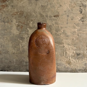 May include: A brown stoneware bottle with a narrow neck and a flat base. The front features a circular embossed stamp with the text "H.J. VAN DER MEULEN" and "SCHIEDAM" surrounding a central monogram, with a smaller oval stamp below the circle.