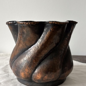 May include: A dark copper-toned metal planter featuring a distinct hammered texture and a spiraling, fluted design. The vessel has a wide, undulating rim with a visible copper-colored edge, showcasing a deep, weathered patina throughout the body.