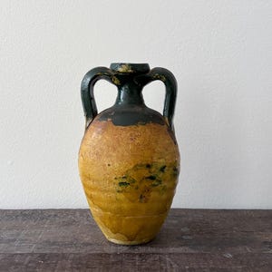 Pode incluir: Um recipiente de cerâmica com duas alças, corpo bulboso e pescoço estreito. A peça tem uma base texturizada de cor amarelo-mostarda com um esmalte verde escuro que escorre da borda e das alças para a parte superior do corpo.