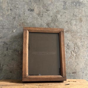 May include: A rectangular wooden picture frame with a natural brown finish and a dark, solid-coloured backing. The frame features mitred corners and a simple, flat profile, enclosing a dark, opaque centre panel with a horizontal crease across the middle.