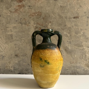 Op de afbeelding: Een keramische vaas in amfoorstijl met een bolvormig lichaam, voorzien van een textuurrijke gele en okerkleurige glazuurlaag met groene vlekken. De hals en de twee gebogen handvatten hebben een donkergroene, glanzende glazuurlaag voor contrast.