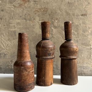 Può includere: Tre forme per cappelli in legno vintage con una finitura marrone naturale. Gli oggetti presentano basi cilindriche e cime affusolate, mostrando segni di usura e una texture venata del legno. Questi strumenti sono disposti in fila.
