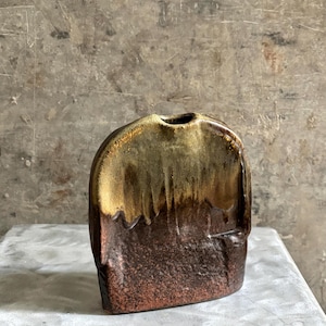 May include: A flat, rounded ceramic vase featuring a two-tone glaze. The upper portion has a metallic gold finish that drips down into a textured, dark brown base. The vessel has a small, centred circular opening at the top and a wide, flat body.