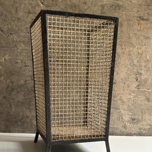 Könnte beinhalten: Ein hoher, rechteckiger Papierkorb mit einem schwarzen Metallrahmen und einem hellbeigen, gewebten Drahtgitterkörper. Der Behälter steht auf vier kurzen, geraden Metallbeinen und verfügt über eine offene Oberseite mit Gittermuster.
